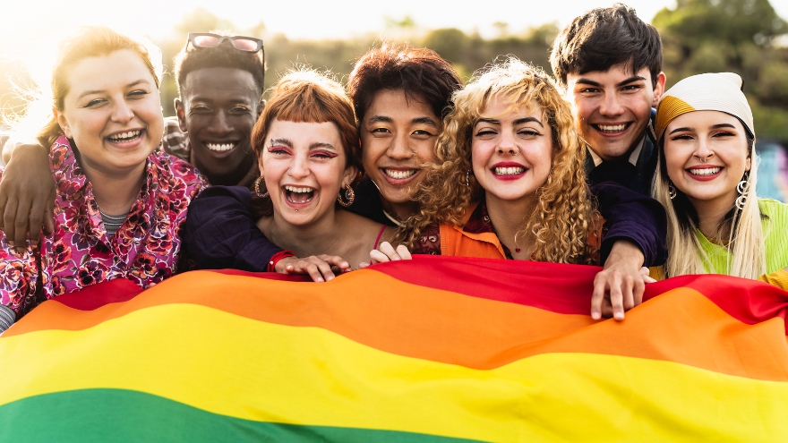 A group of teens smile behind the LGBTQIA+ pride flag.