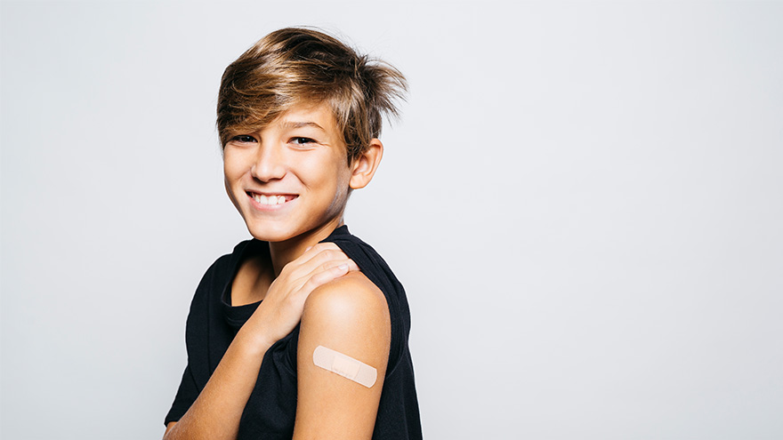 boy with blonde hair smiling at camera showing bandaid on his upper arm from vaccine